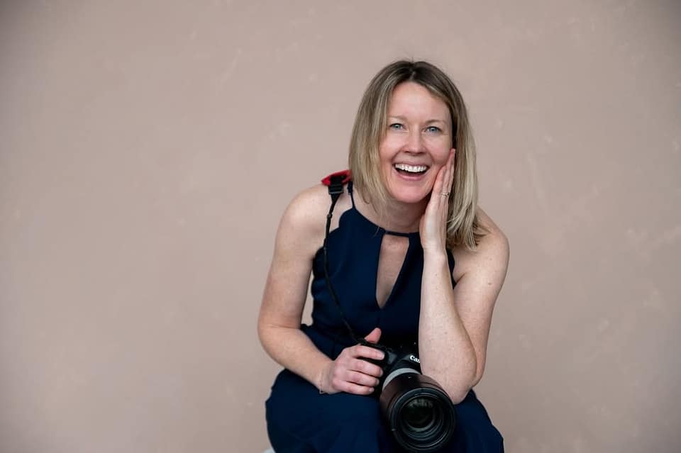 Calgary Wedding Photographer Shannon Hutchison holds her camera and smiles happily at the camera, wide view